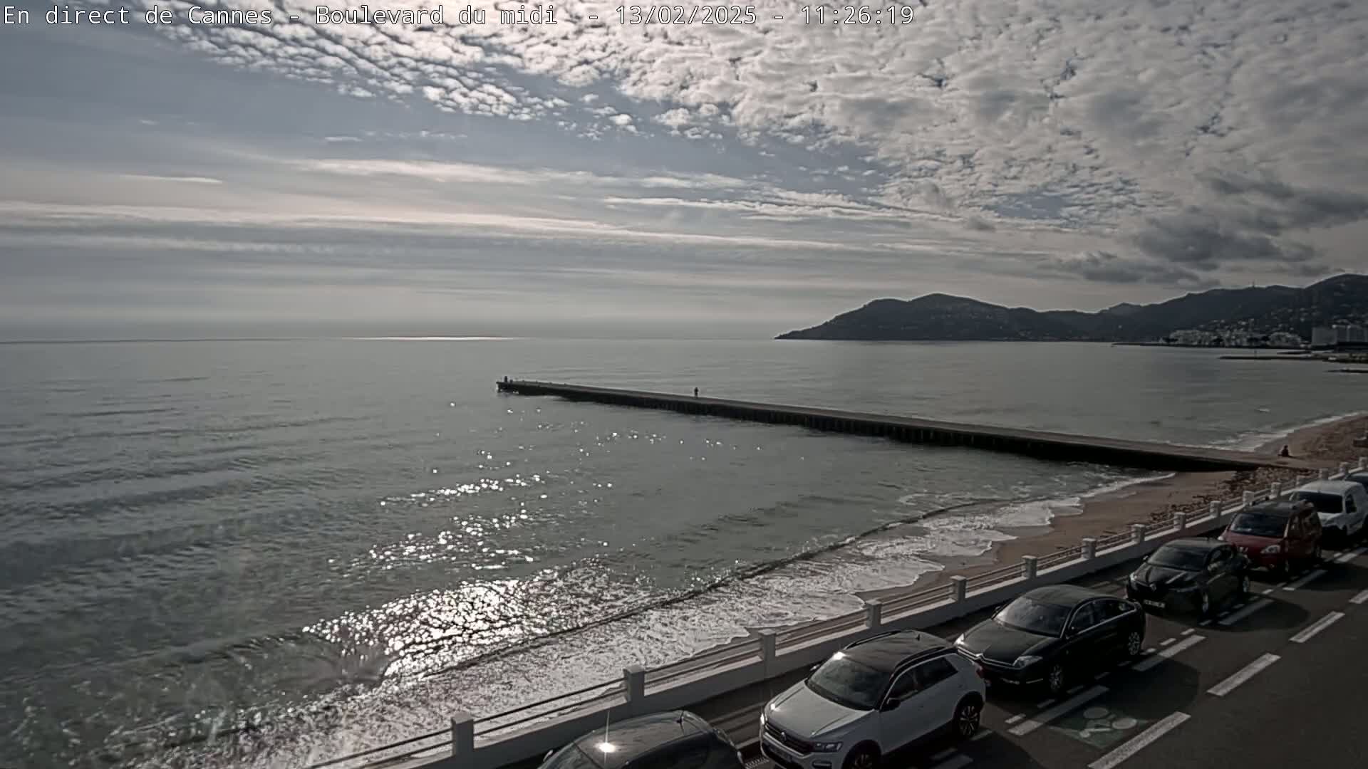 A long pier extends into a calm sea under a partly cloudy sky, with cars parked along a seaside road and a sandy beach in the foreground.