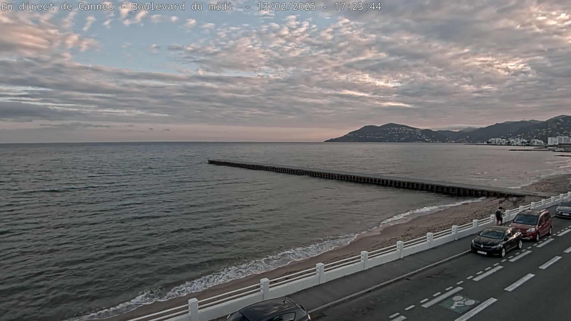 A long breakwater stretches into a calm sea under a cloudy sky, with a sandy beach, cars parked along a road, and distant hills visible.