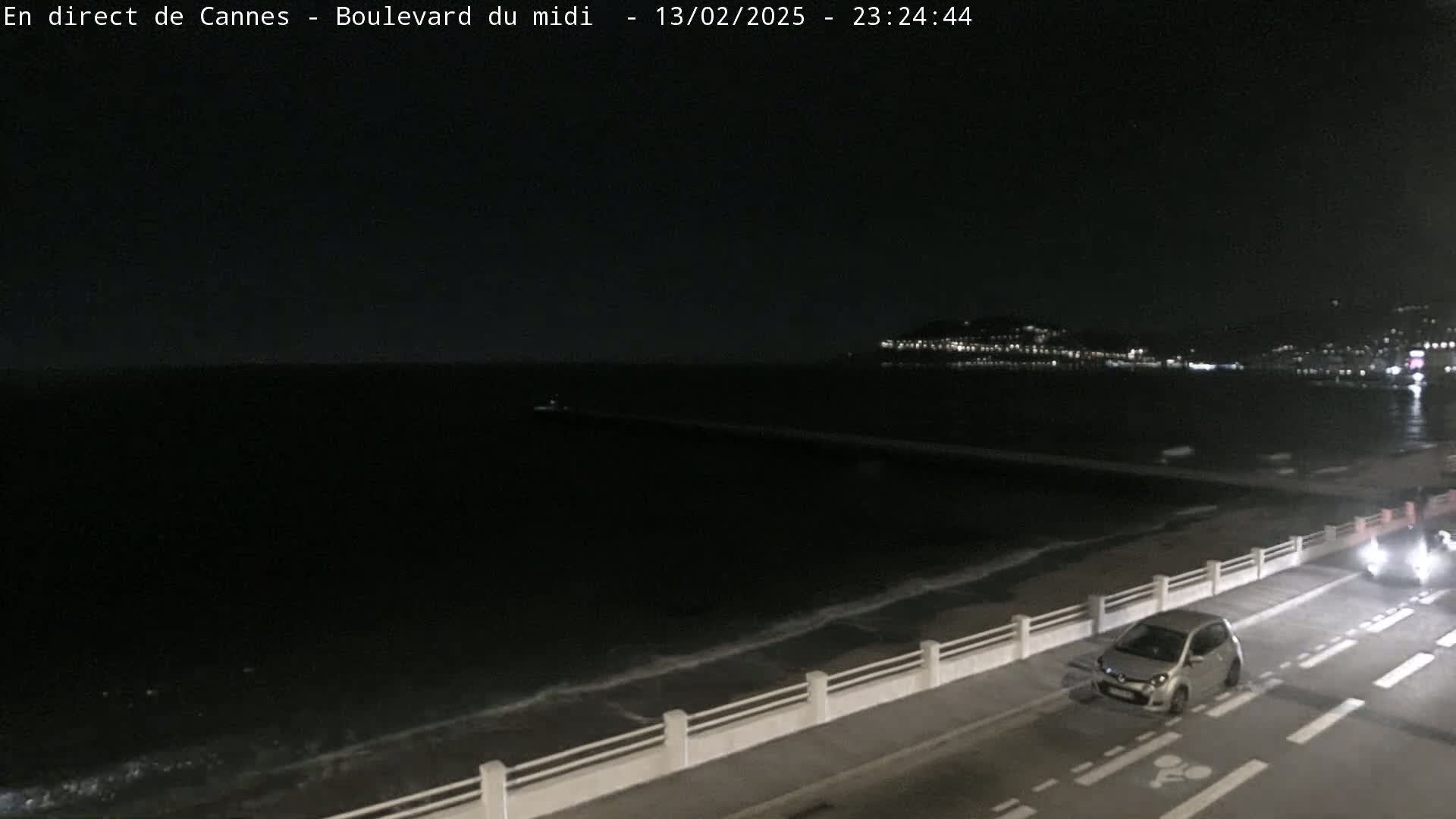 A nighttime coastal scene shows a dark ocean with gentle waves lapping a seawall next to a road where a car is parked.