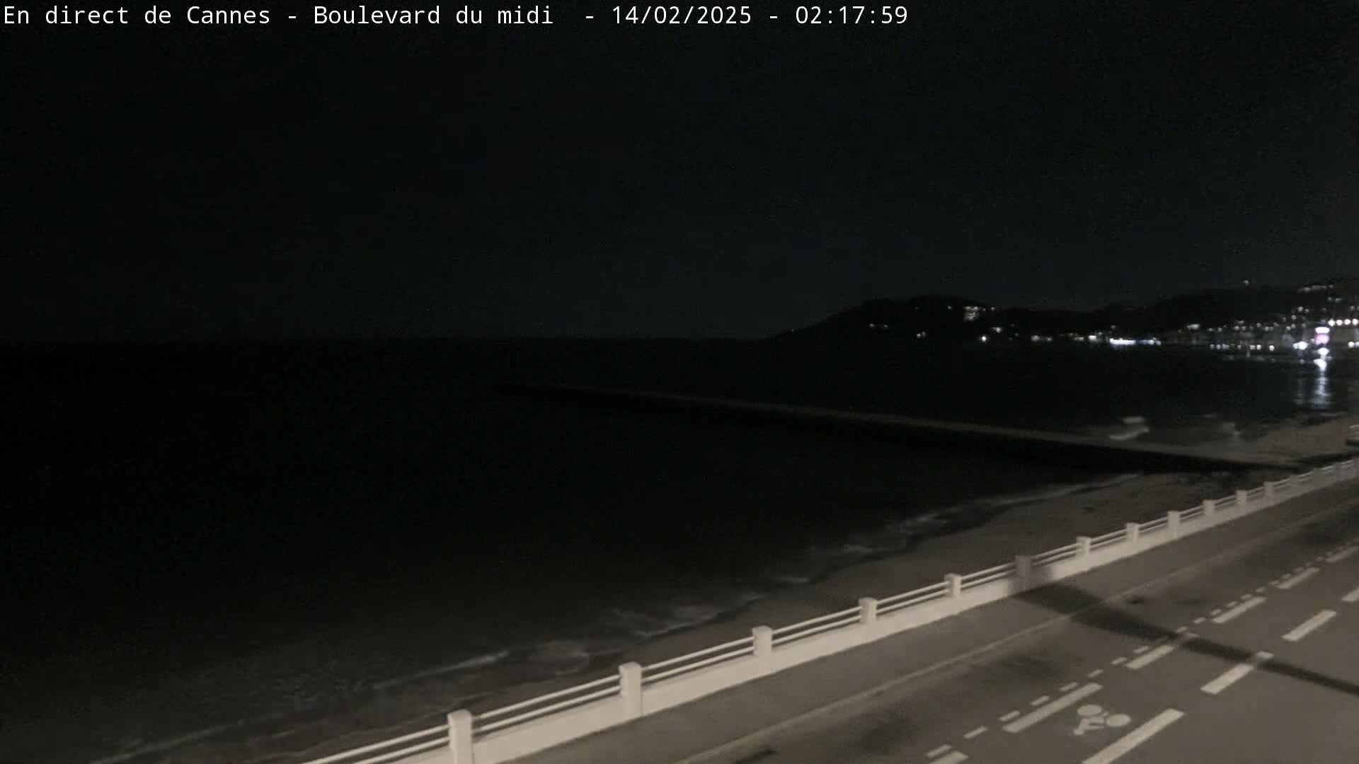 A nighttime coastal scene shows a dark sea with gentle waves lapping a sandy shore, a low seawall, and a roadway with bike lanes.