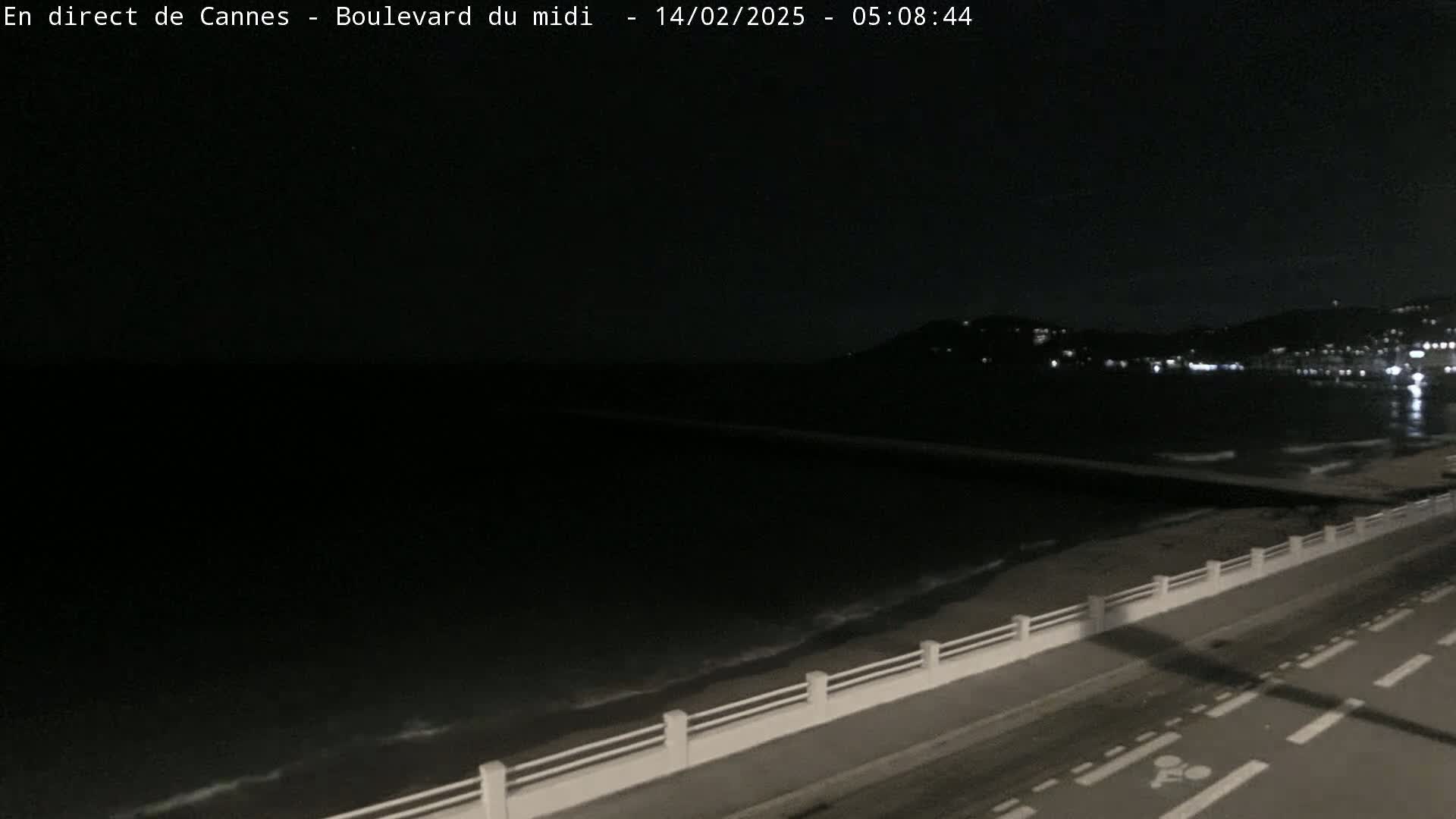 A dark, nighttime coastal scene shows a gently waving sea lapping a beach with a road and low white railing running along the shore, while lights from a distant city glimmer across the water.