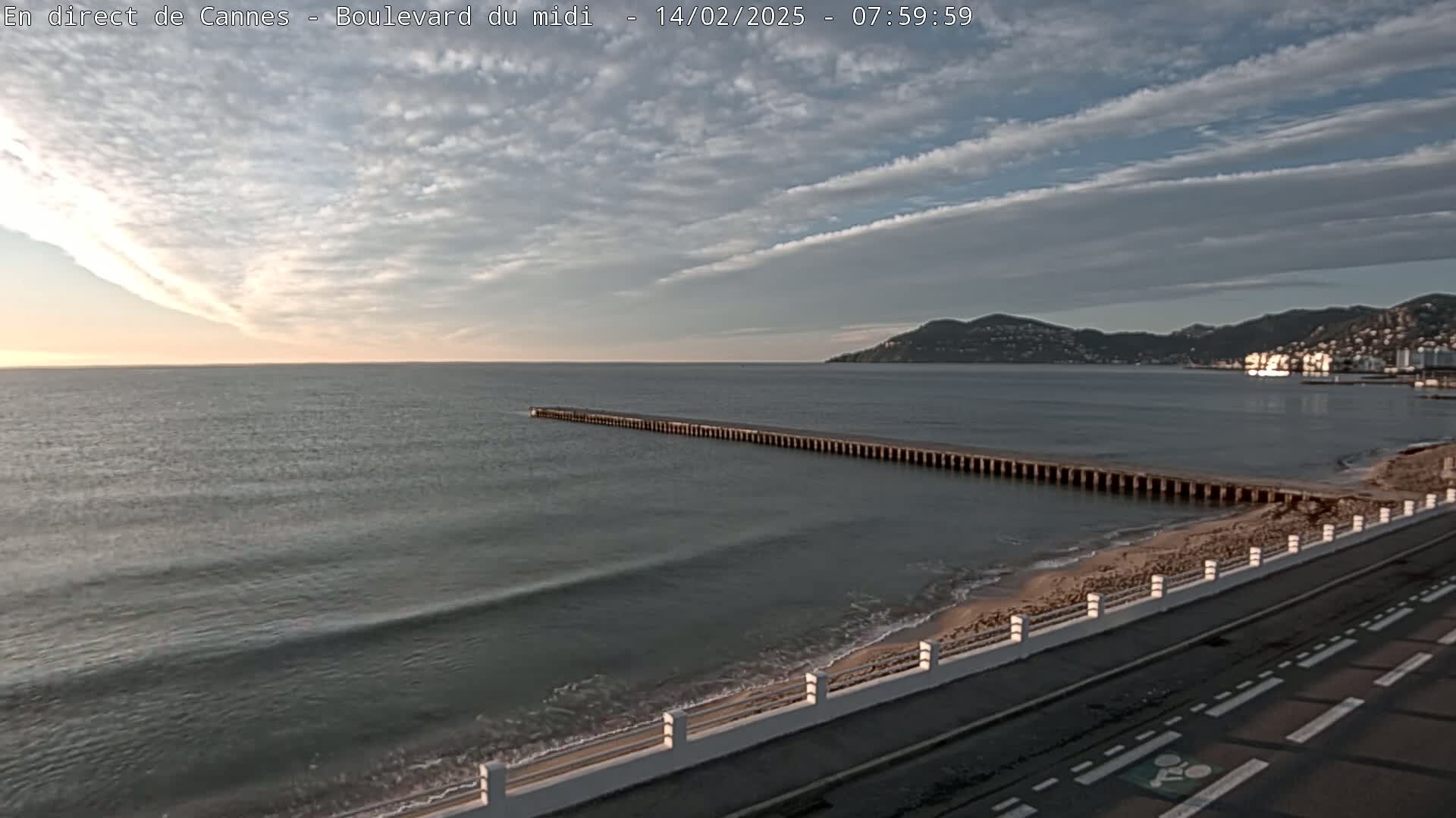 A long pier extends into a calm sea under a partly cloudy sky, with a sandy beach and road visible in the foreground and distant hills in the background.