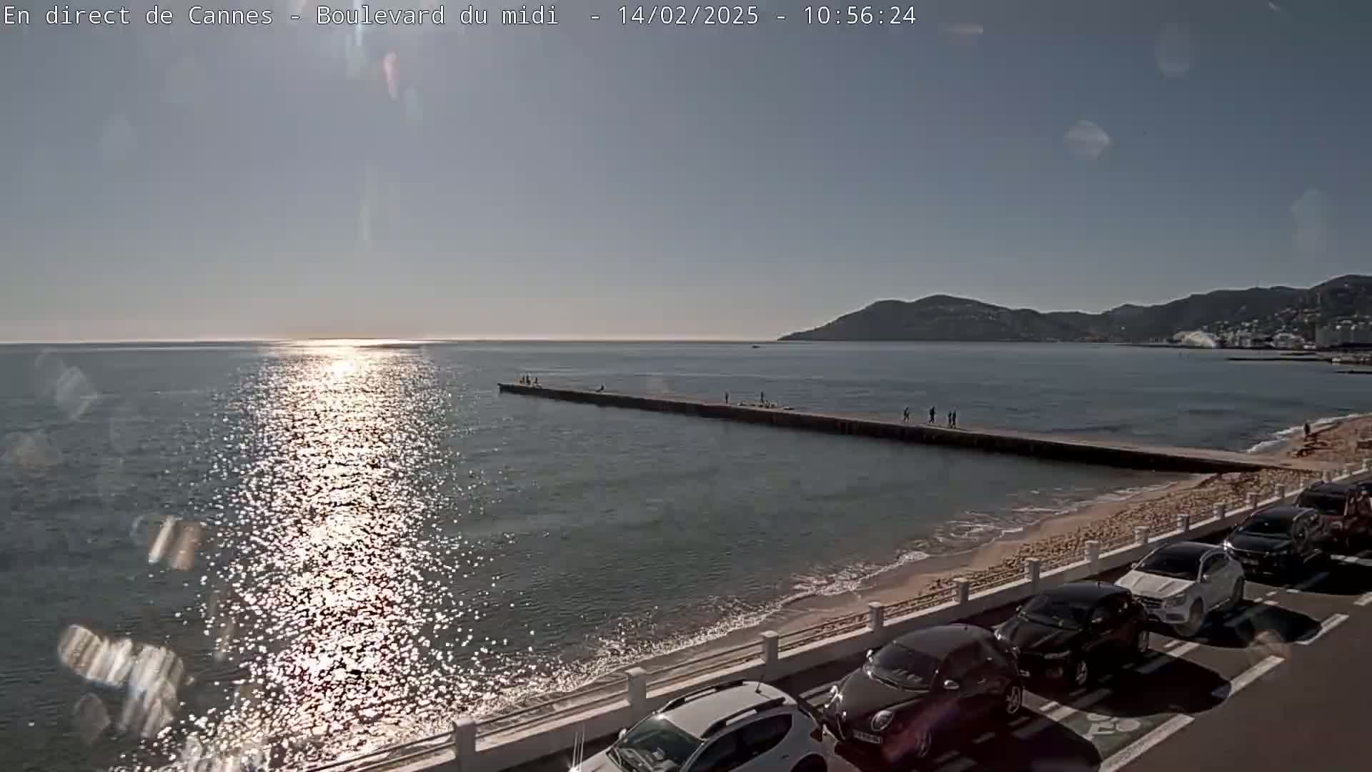 A long pier extends into a calm, sun-lit sea near a sandy beach lined with parked cars under a clear sky.