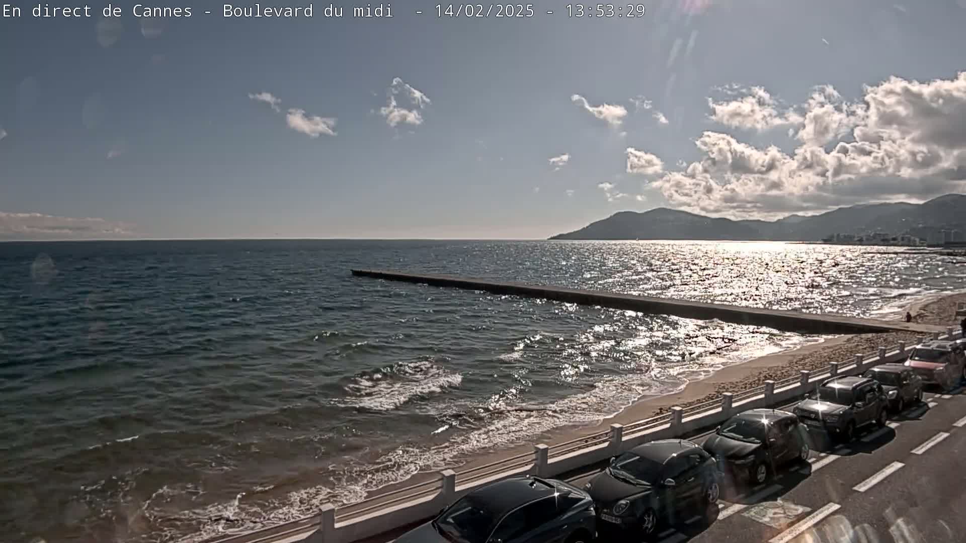 A partly sunny day shows a beach with parked cars along a seawall, a long jetty extending into a somewhat choppy sea, and mountains in the distance.