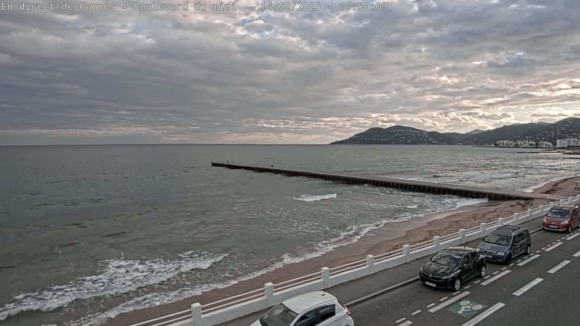 A long pier extends into a gray, wavy sea under a cloudy sky, with a sandy beach, a white railing, and several cars parked along a road in the foreground.