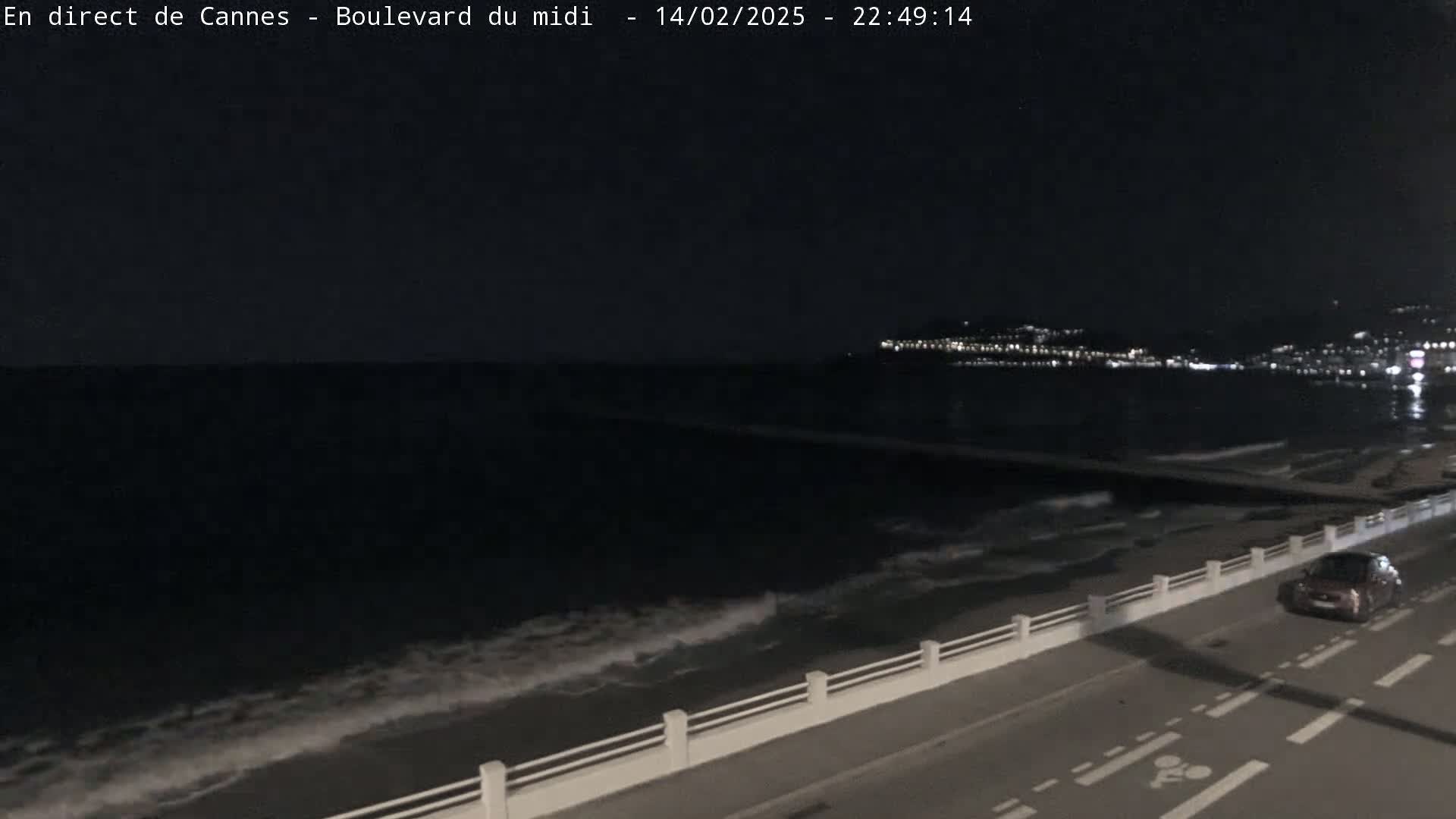 A dark, nighttime coastal scene shows a road next to a sea with small waves, a single car on the road, and a city illuminated in the distance.