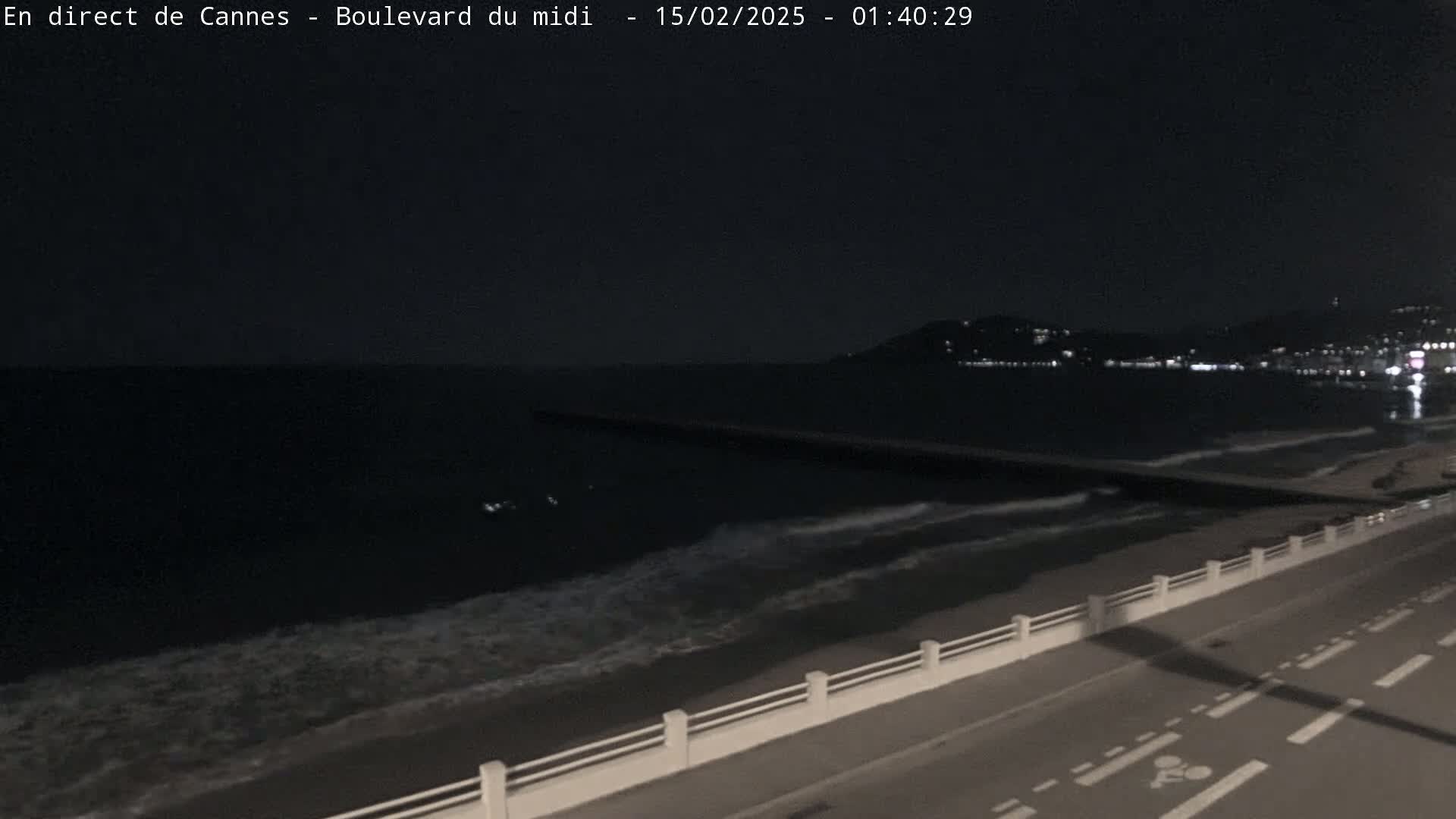 A dark nighttime coastal scene shows a calm sea with small waves lapping a shoreline, a long breakwater extending into the water, and a road with a low white railing running alongside.