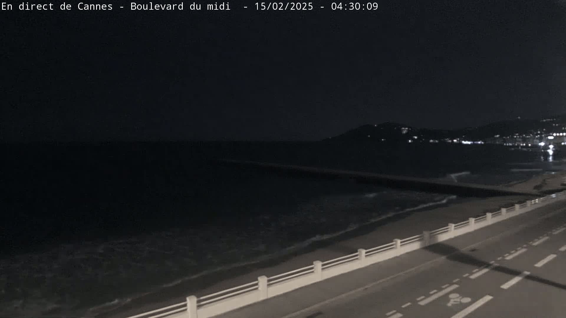 A nighttime coastal scene shows dark water with gentle waves lapping a shoreline with a road and low wall, and distant lights illuminate a hillside in the background.