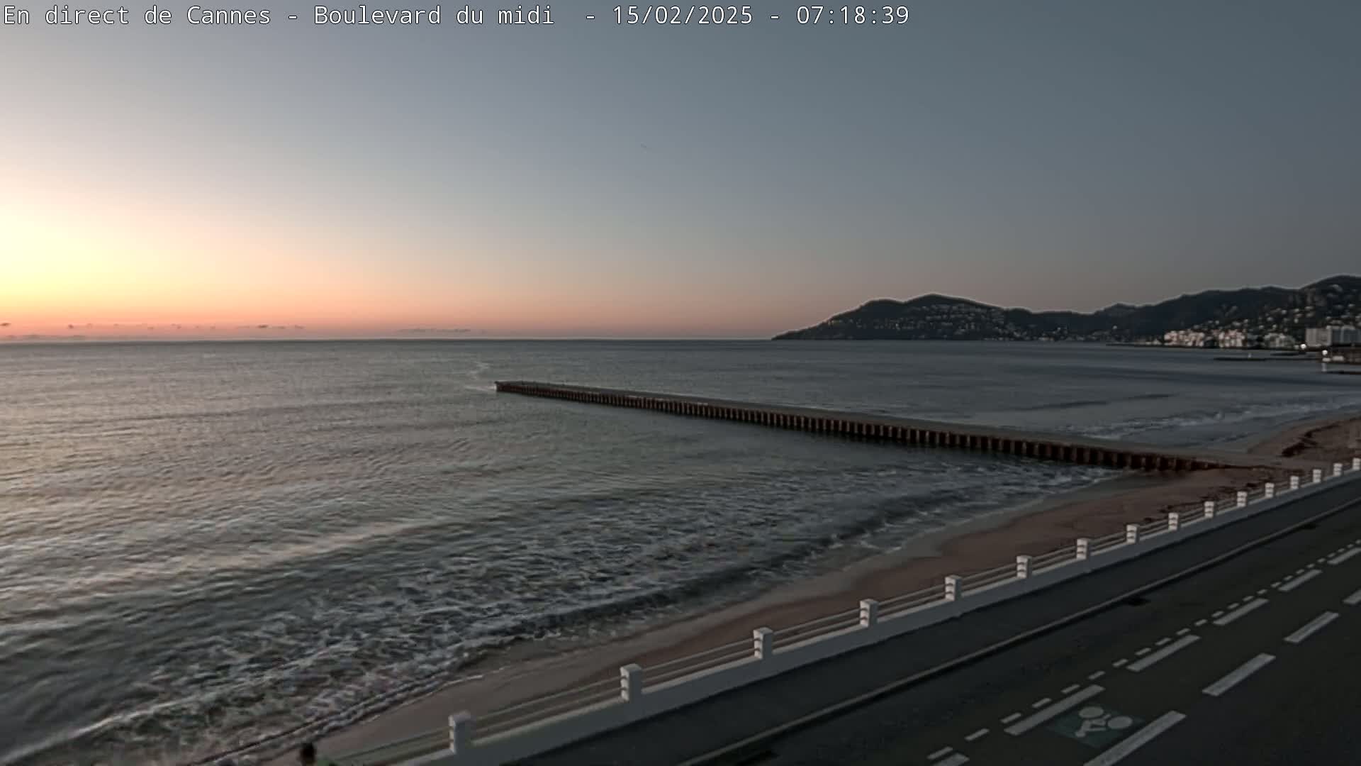 A long pier extends into a calm sea during a sunrise, with a road and low wall separating the beach from the roadway.