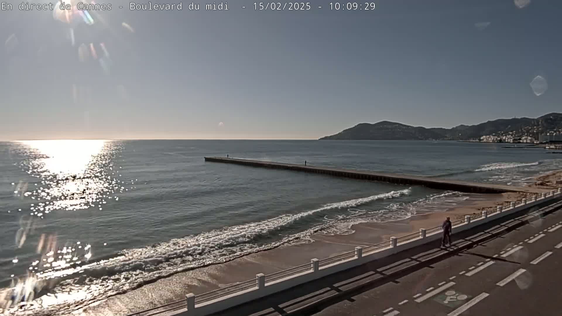A long pier extends into a calm, sunlit ocean with gentle waves lapping a sandy beach, and a person walks along a paved path next to the water under a clear sky.