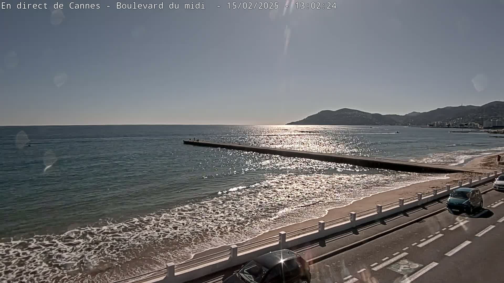 A long pier extends into a calm, sunlit sea with gentle waves lapping a sandy beach, backed by a road and distant hills under a clear sky.