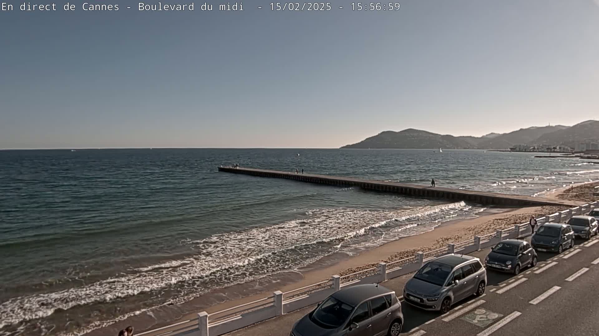 A long pier extends into a calm sea with small waves lapping a sandy beach lined with parked cars under a clear, sunny sky.