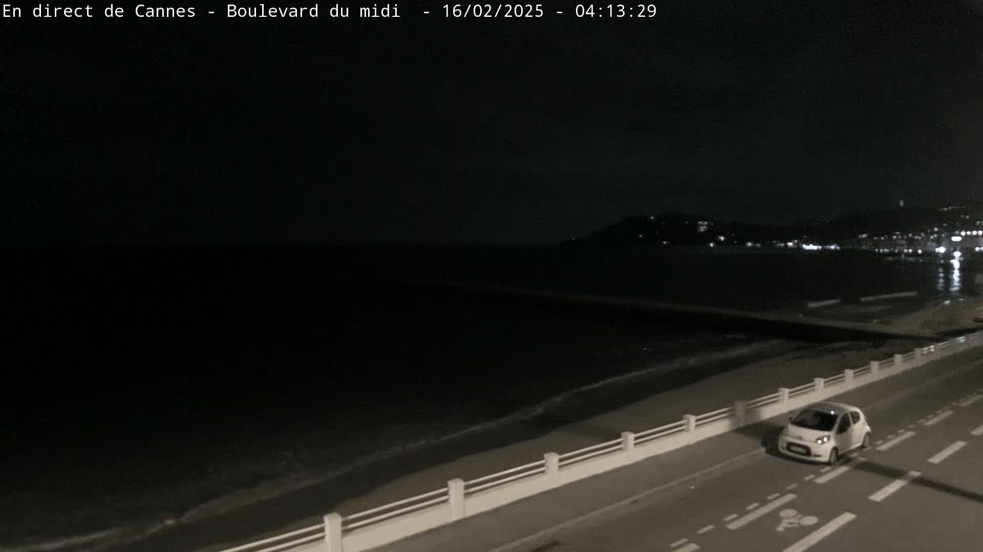 A white car drives along a coastal road at night, with dark water and a distant, dimly lit cityscape visible under a cloudy sky.