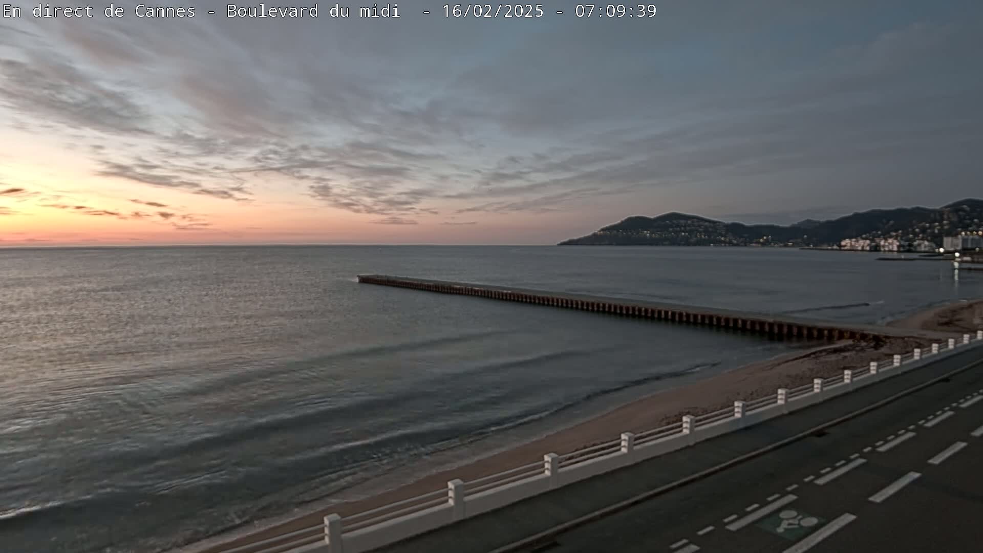 A long pier extends into a calm sea at sunset, with a paved road and low wall bordering a sandy beach and hills visible in the distance.