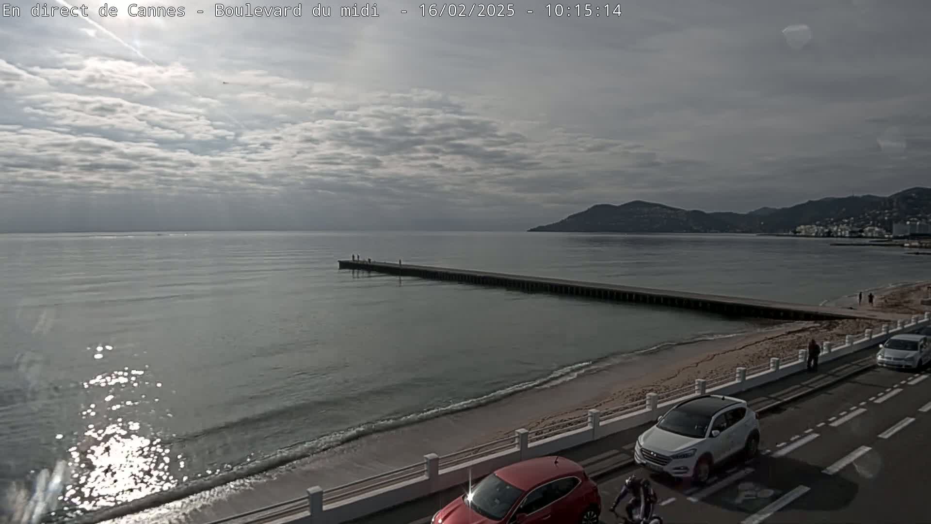 A long pier extends into a calm sea under a cloudy sky, with cars parked along a coastal road next to a sandy beach.