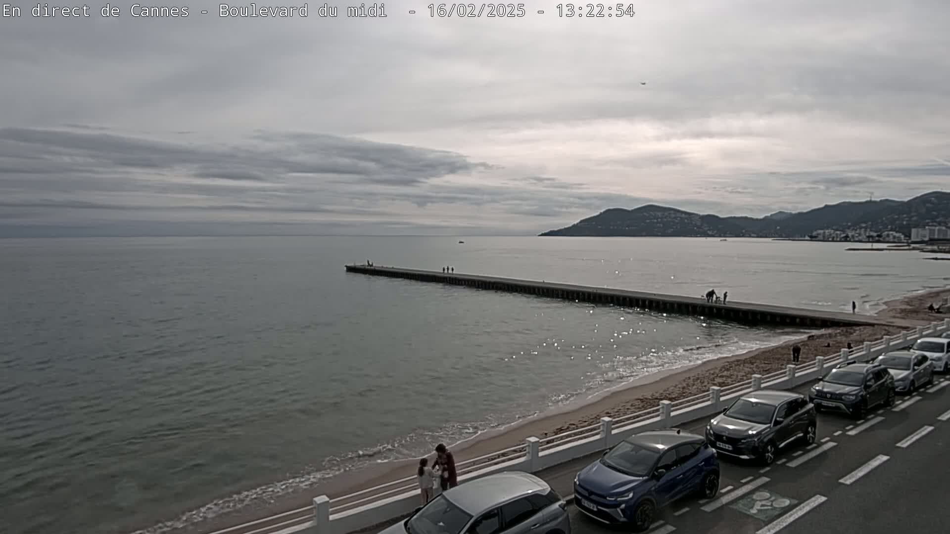 A long pier extends into a calm, gray sea under a cloudy sky, with a sandy beach and parked cars lining a road in the foreground.