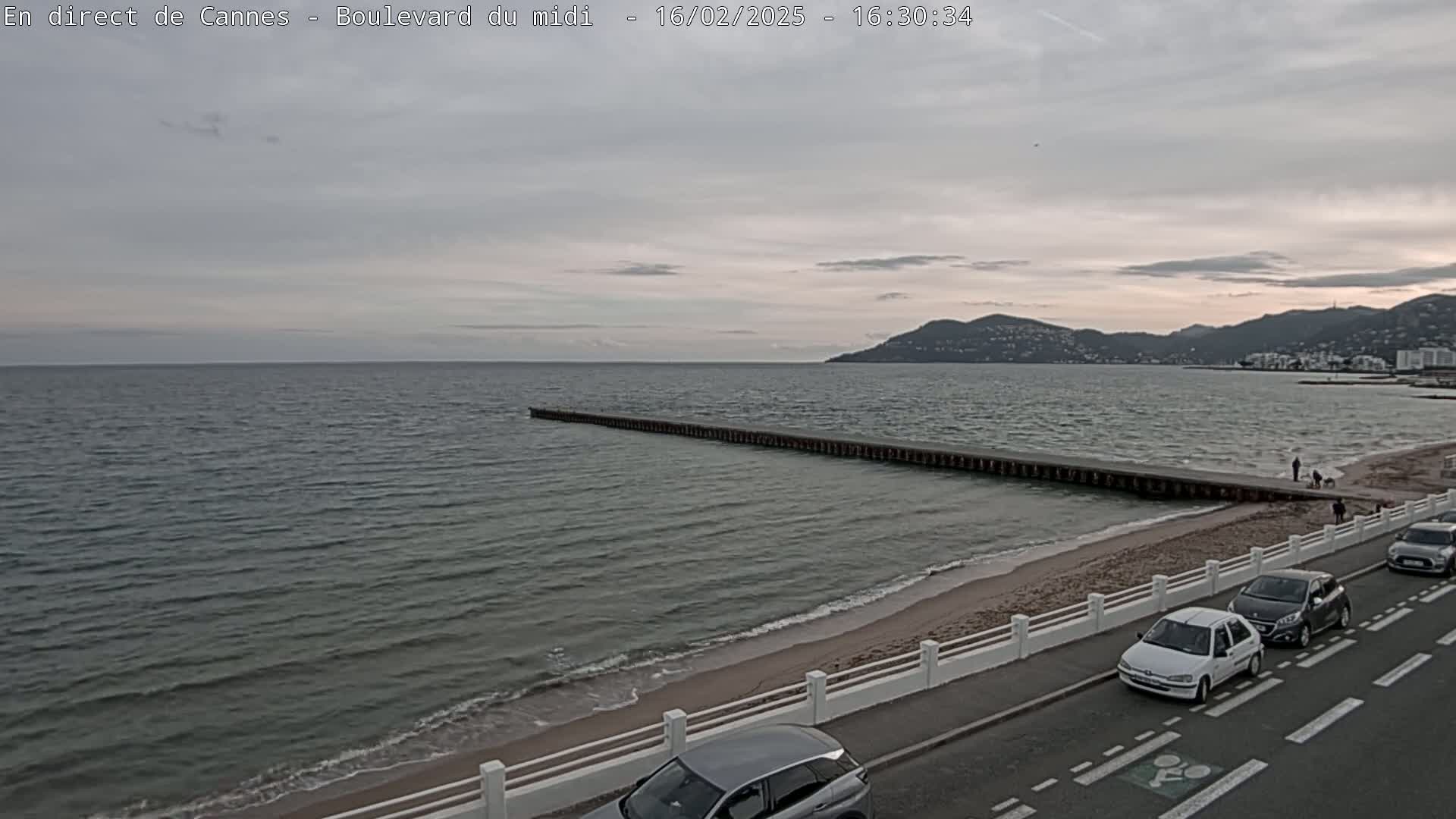 A mostly cloudy evening scene shows a long breakwater extending from a sandy beach into a calm sea, with several cars parked along a roadside and a few people walking on the beach.