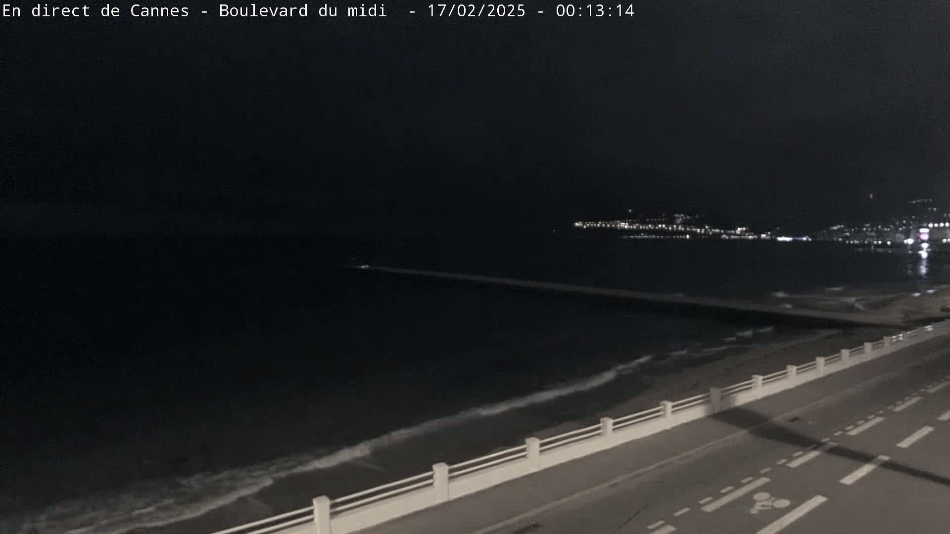 A nighttime coastal scene shows dark water lapping at a shore with a roadway and low white wall, distant city lights visible across the water.