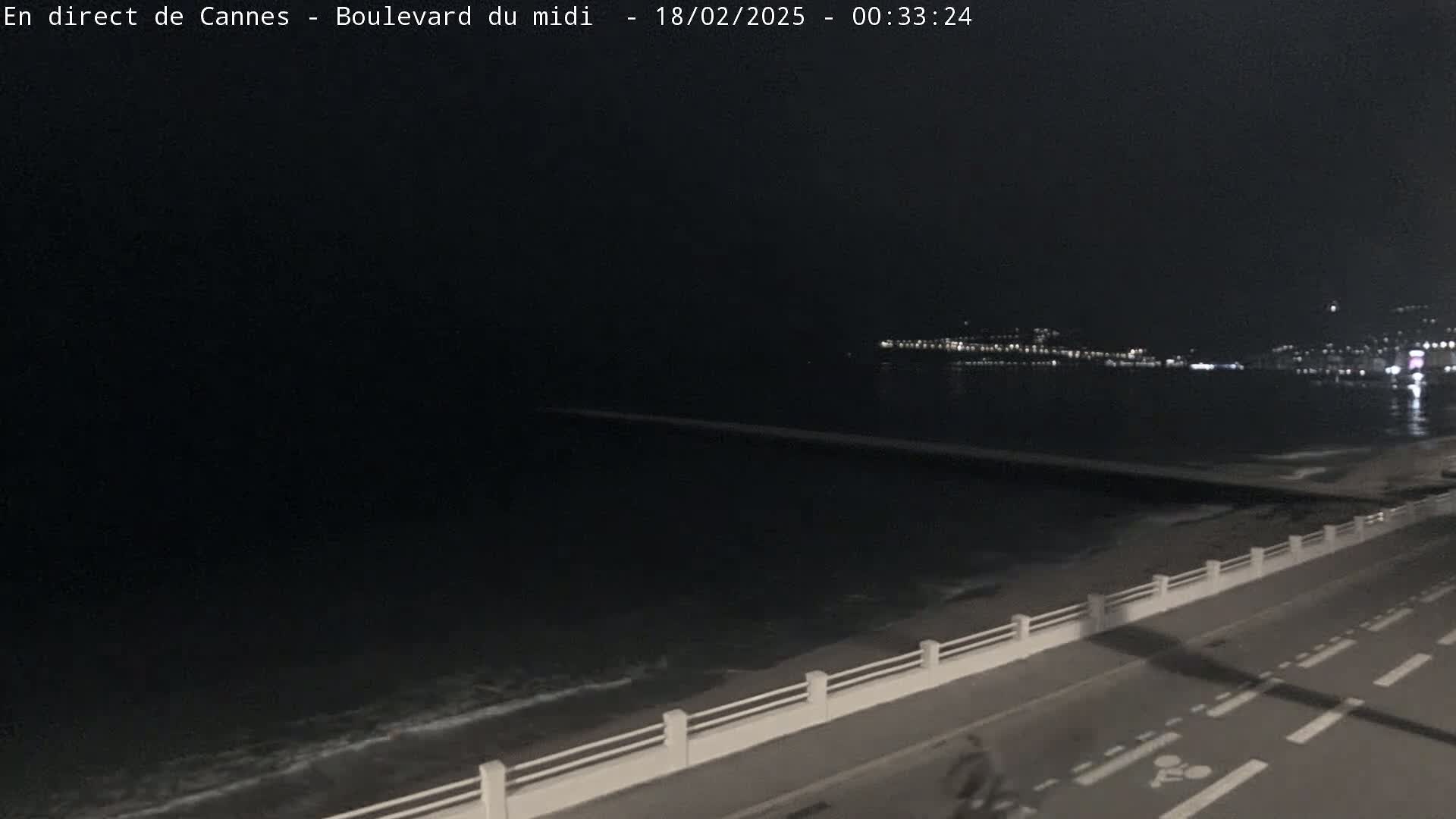 A nighttime view of a dark ocean with a city skyline faintly visible in the distance, a long breakwater stretching into the water, and a road with a bike lane next to a low seawall.
