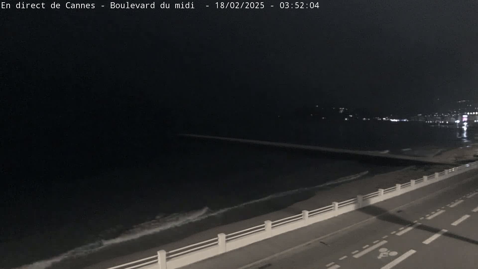 A nighttime view of a dark ocean with gentle waves lapping a shoreline, a long breakwater extending into the water, and a road with a dividing barrier in the foreground.