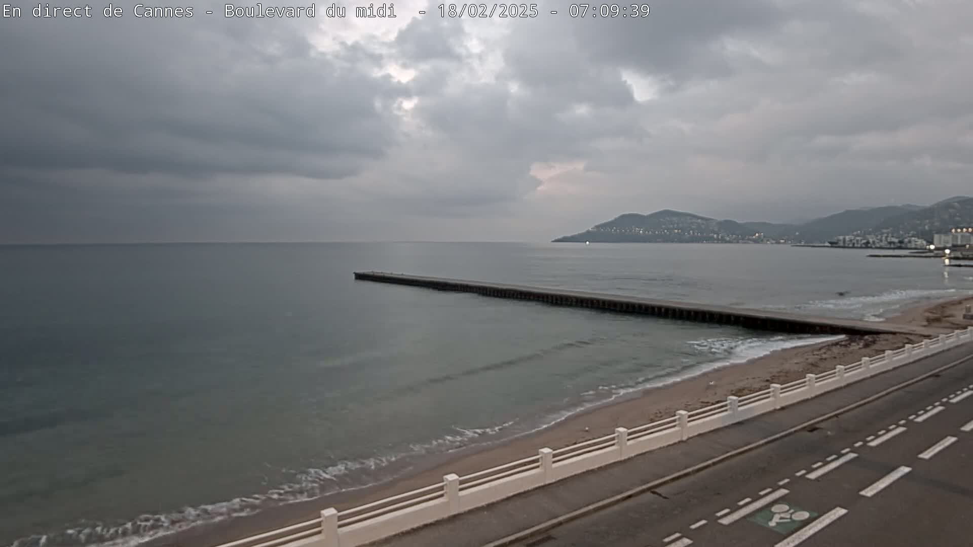 A long pier extends into a calm, gray sea under an overcast sky, with a sandy beach and road visible in the foreground and distant hills in the background.