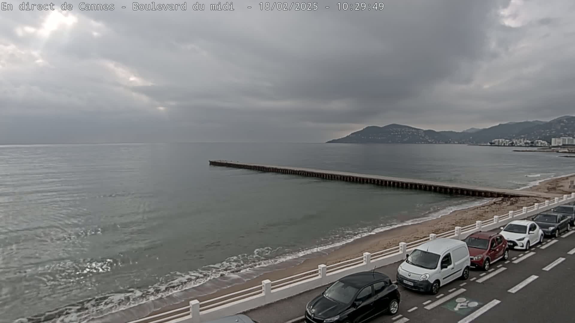 A long pier extends into a calm, grey sea under an overcast sky, with several cars parked along a sandy beach.