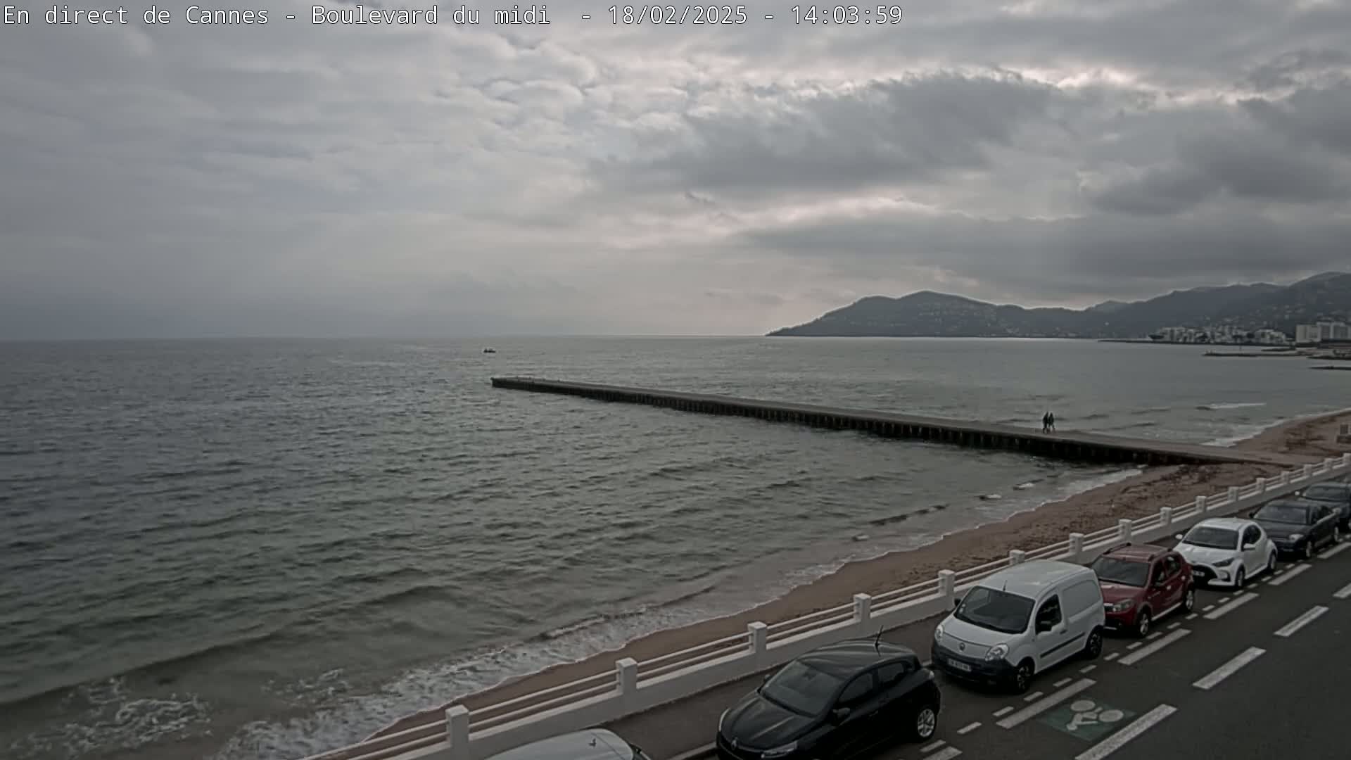 A long pier extends into a gray, somewhat choppy sea under a cloudy sky, with a sandy beach and parked cars lining a road in the foreground.