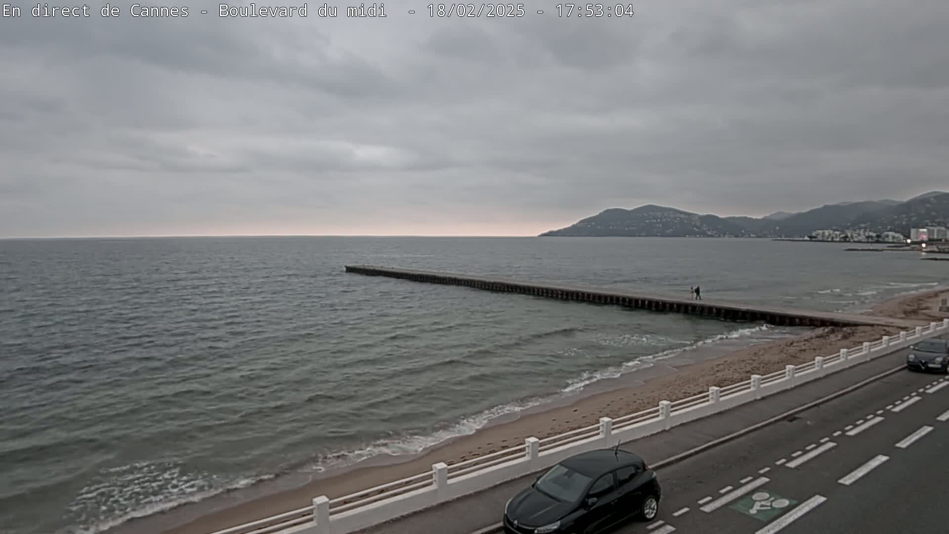 A mostly cloudy day at a beach shows a long jetty extending into a calm sea, with a couple walking along it and cars parked alongside a road next to the sand.