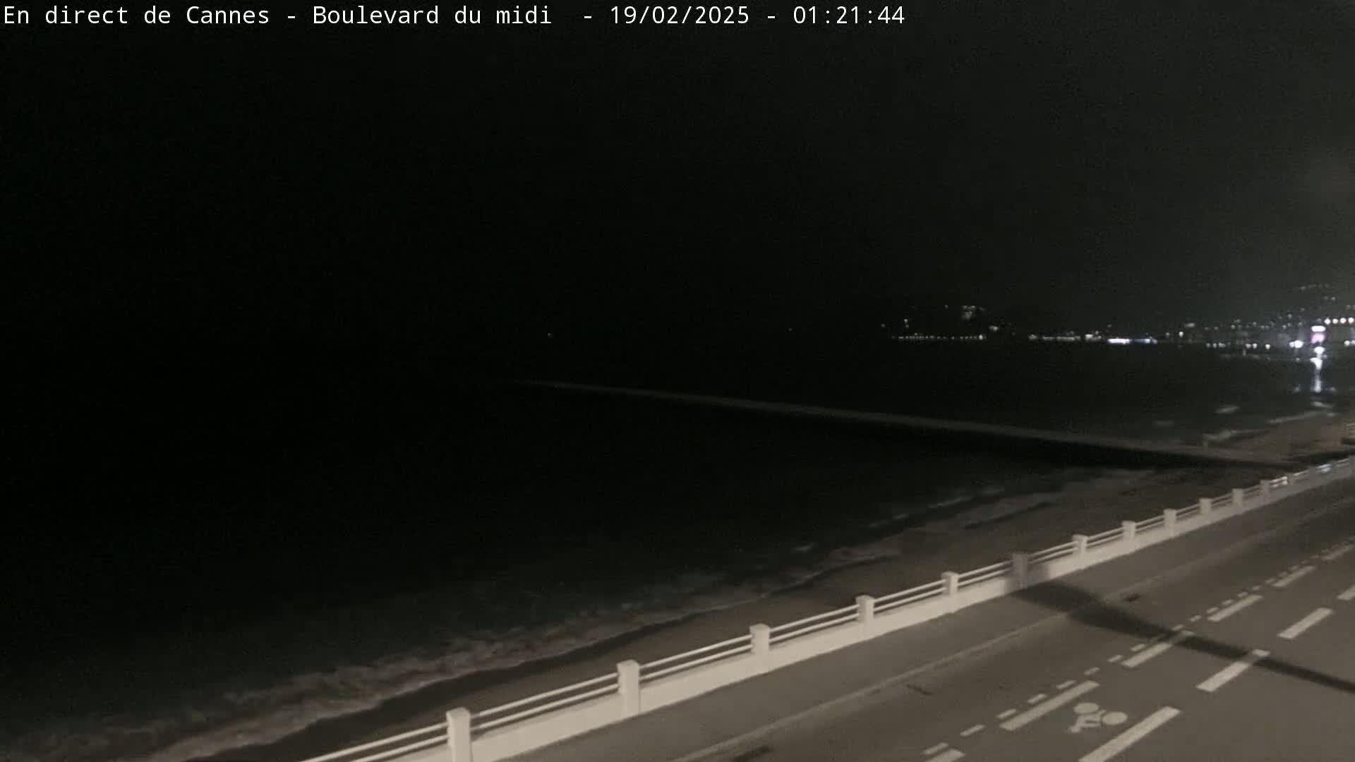 A dark, nighttime coastal scene shows a paved road next to a low wall separating it from a gently lapping ocean, with lights faintly visible in the distance.