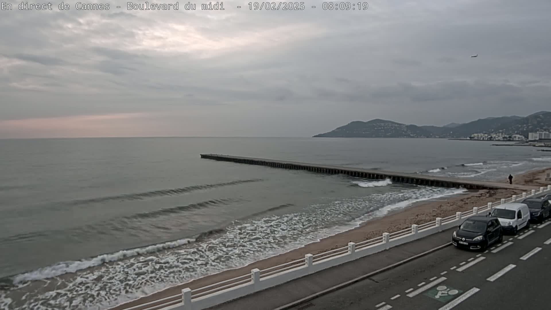 A long pier extends into a gray, wavy sea under an overcast sky, with a sandy beach, a road with parked cars, and distant hills visible in the background.
