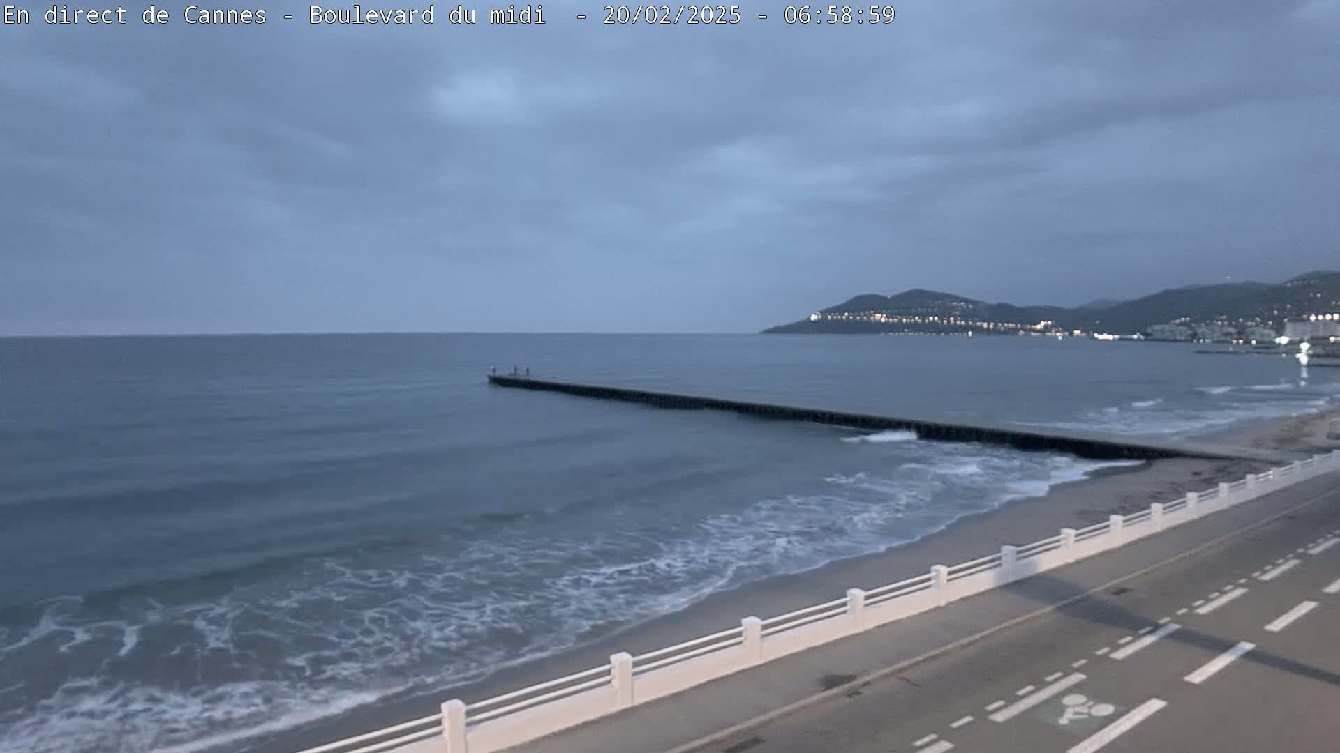 A long pier extends into a calm, gray sea under an overcast sky, next to a road with a white railing separating it from the beach.