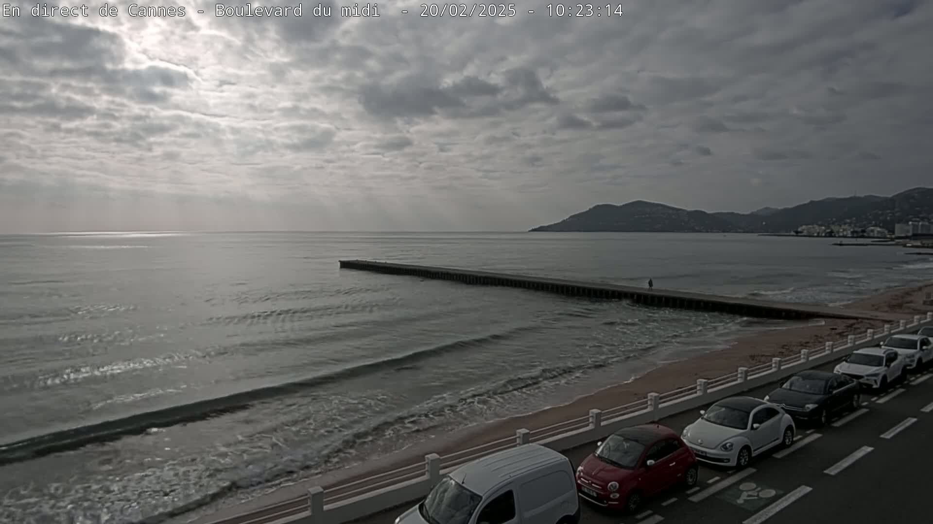A long, low breakwater extends into a grey sea under a mostly cloudy sky, with several cars parked along a sandy beach next to a road.