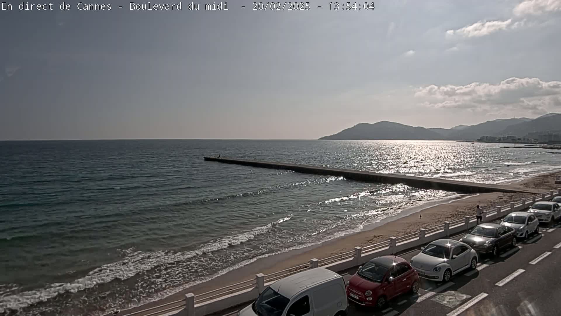 A partly sunny day shows a sandy beach with gently lapping waves, a long jetty extending into the sea, and a line of cars parked along a roadway next to the beach.