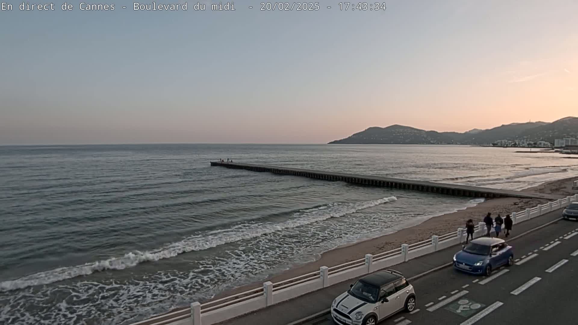 A calm sea with small waves laps a sandy beach at sunset, under a pale sky; a long pier extends into the water, and several people walk along the shore near parked cars.