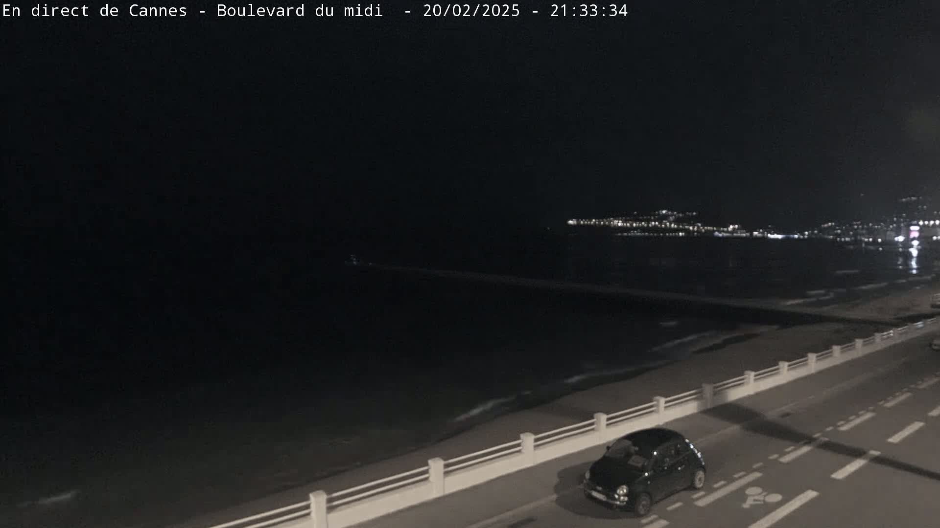 A dark-colored car is parked on a road next to a beach at night, with a city's lights visible in the distance across the water.