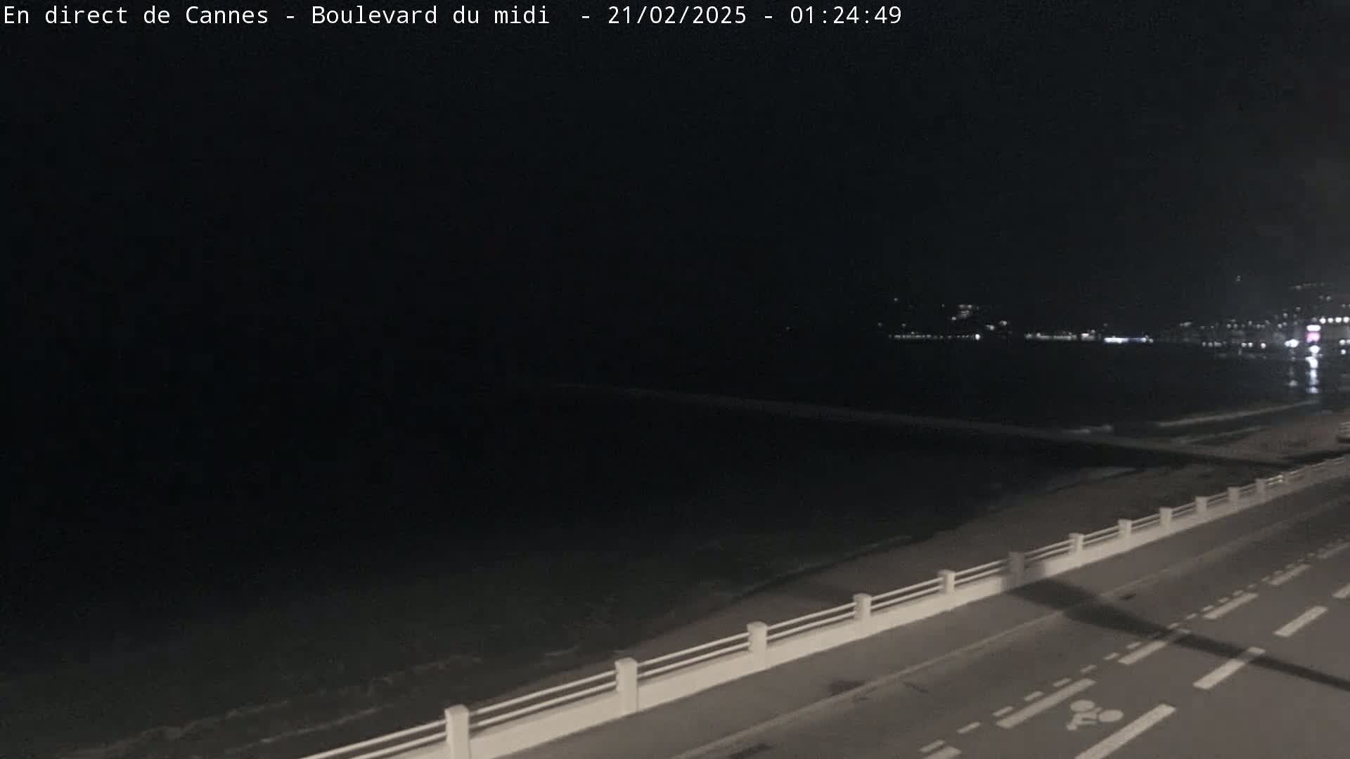 A nighttime, coastal view shows a dark ocean, a beach, and a road with a low wall separating them, under a clear, dark sky.