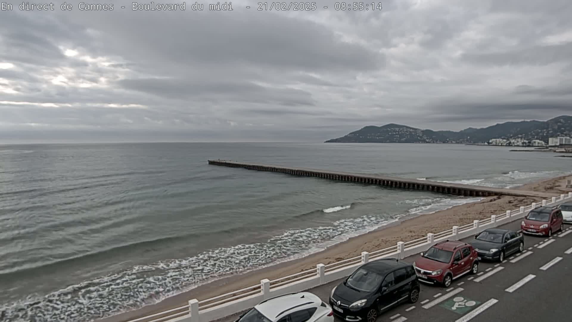 A long pier extends into a calm, gray sea under an overcast sky, with a sandy beach and several cars parked along a roadside in the foreground.