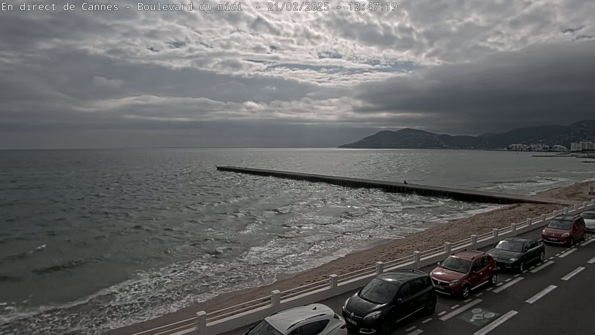 A mostly cloudy day shows a view of a beach with a long jetty extending into a wavy sea, and several cars parked along the roadside.