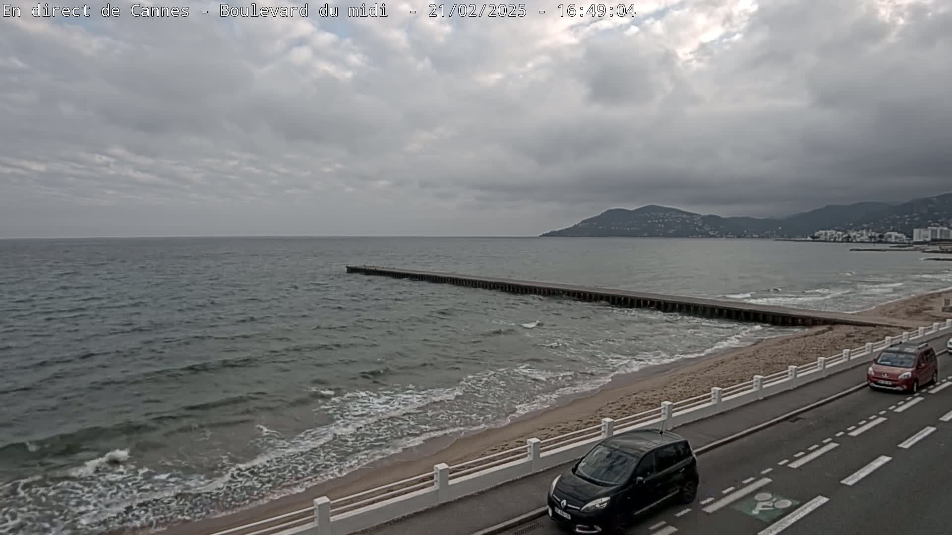 A long pier extends into a wavy sea under a cloudy sky, with a road and two cars visible along a sandy beach and distant hills.