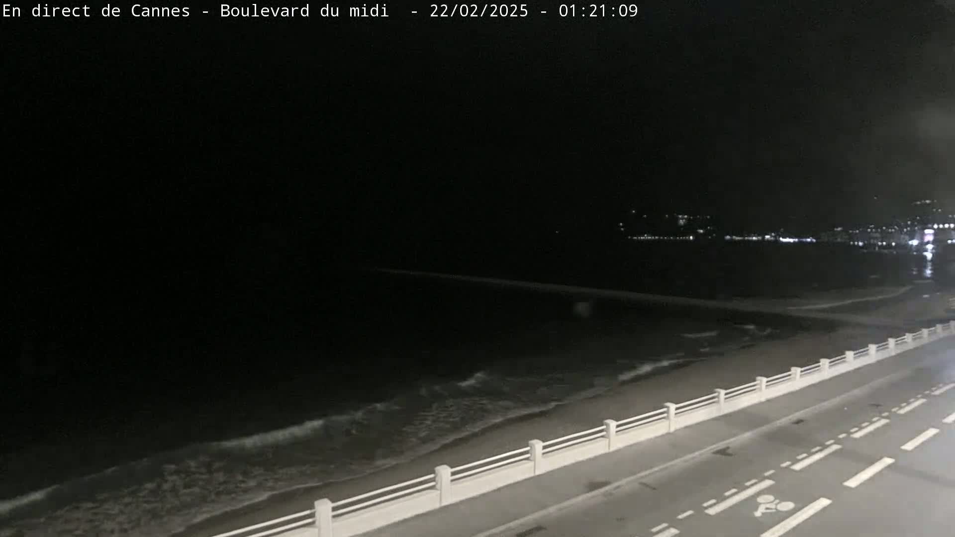 A dark nighttime coastal scene shows a gently lapping ocean with a roadway and low wall separating it from the road.