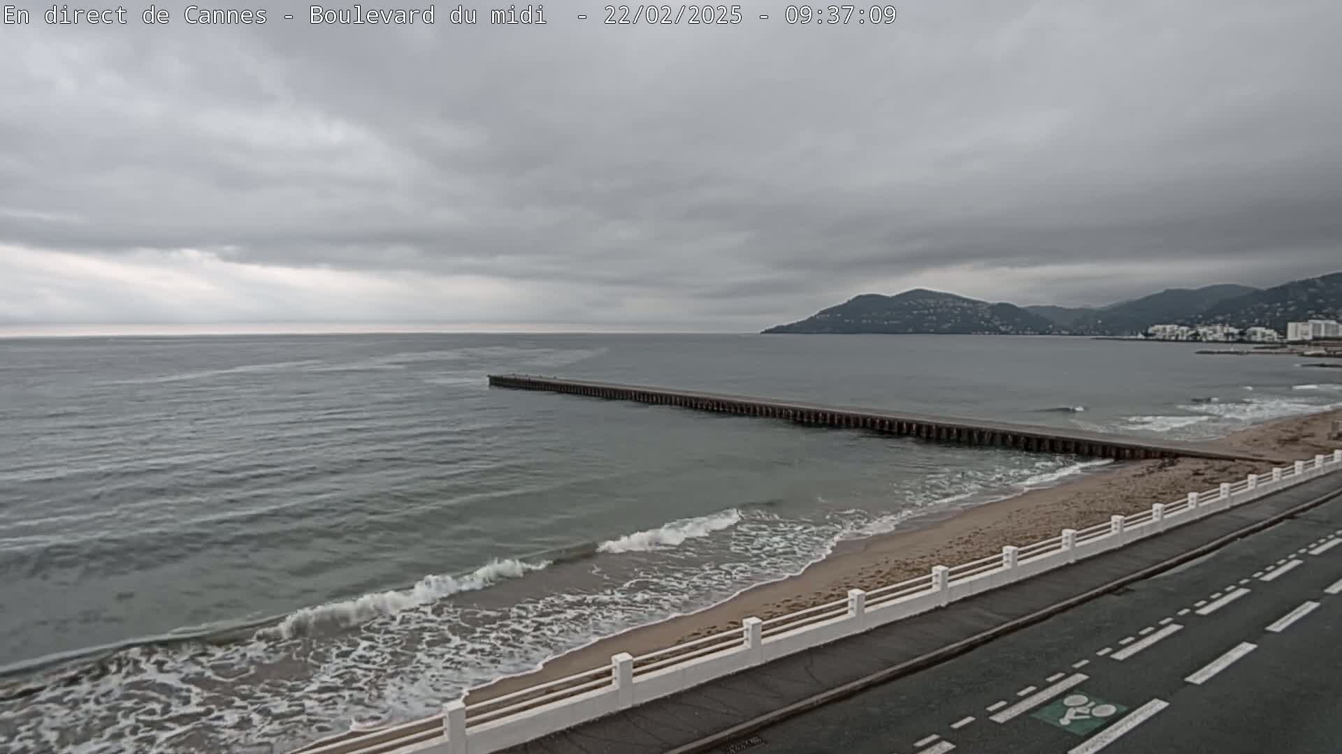 A long pier extends into a calm sea under an overcast sky, with a sandy beach and road visible in the foreground.