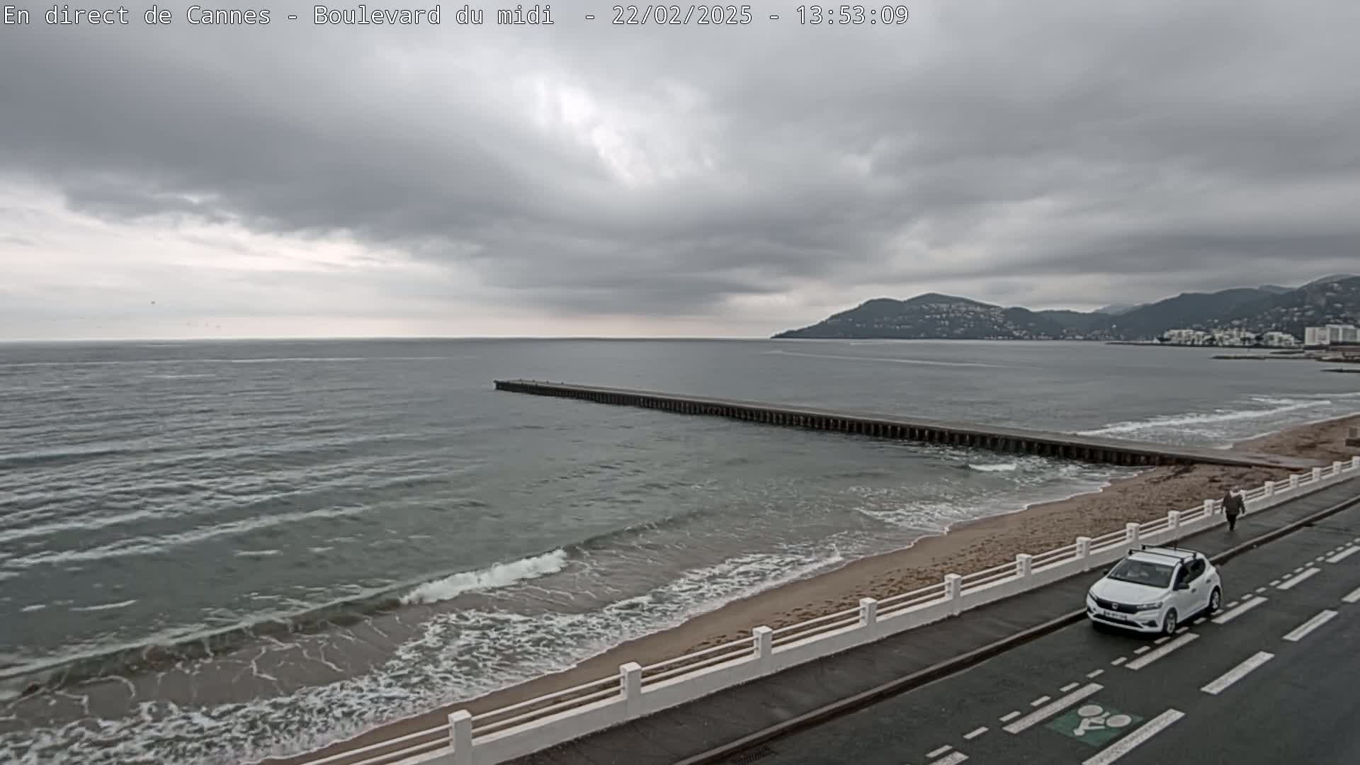 A long pier extends into a grey, wavy ocean under a cloudy sky, with a sandy beach and a white car on a nearby road.