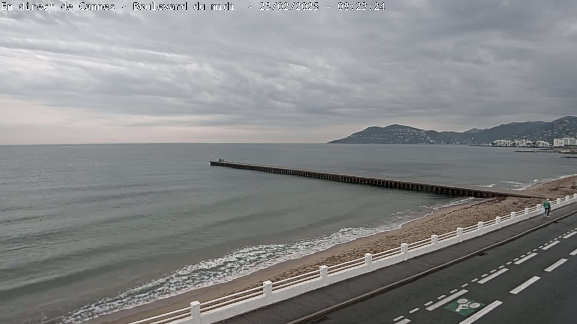A long pier extends into a calm, gray sea under a cloudy sky, with a sandy beach and road alongside it.