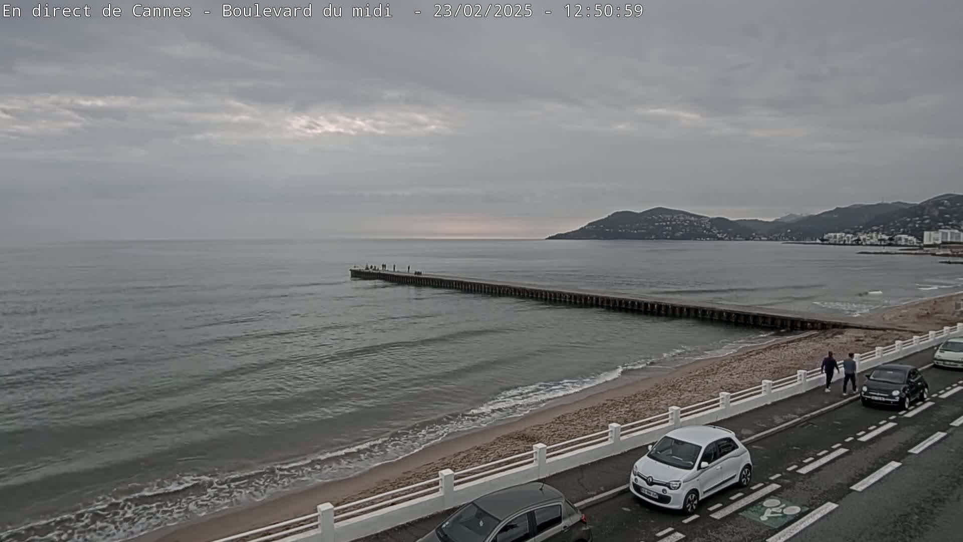 A long pier extends into a calm, gray sea under an overcast sky, with a sandy beach, a road, and several cars visible along the shore.