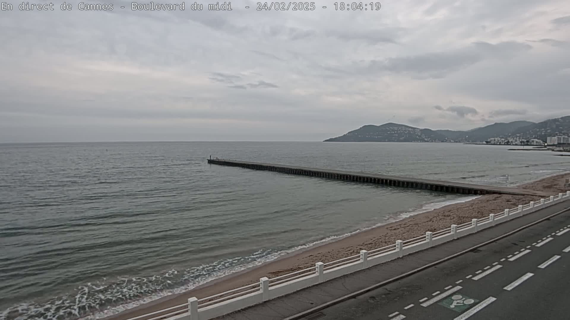 A long pier extends into a calm, grey sea under a cloudy sky, alongside a sandy beach and road.