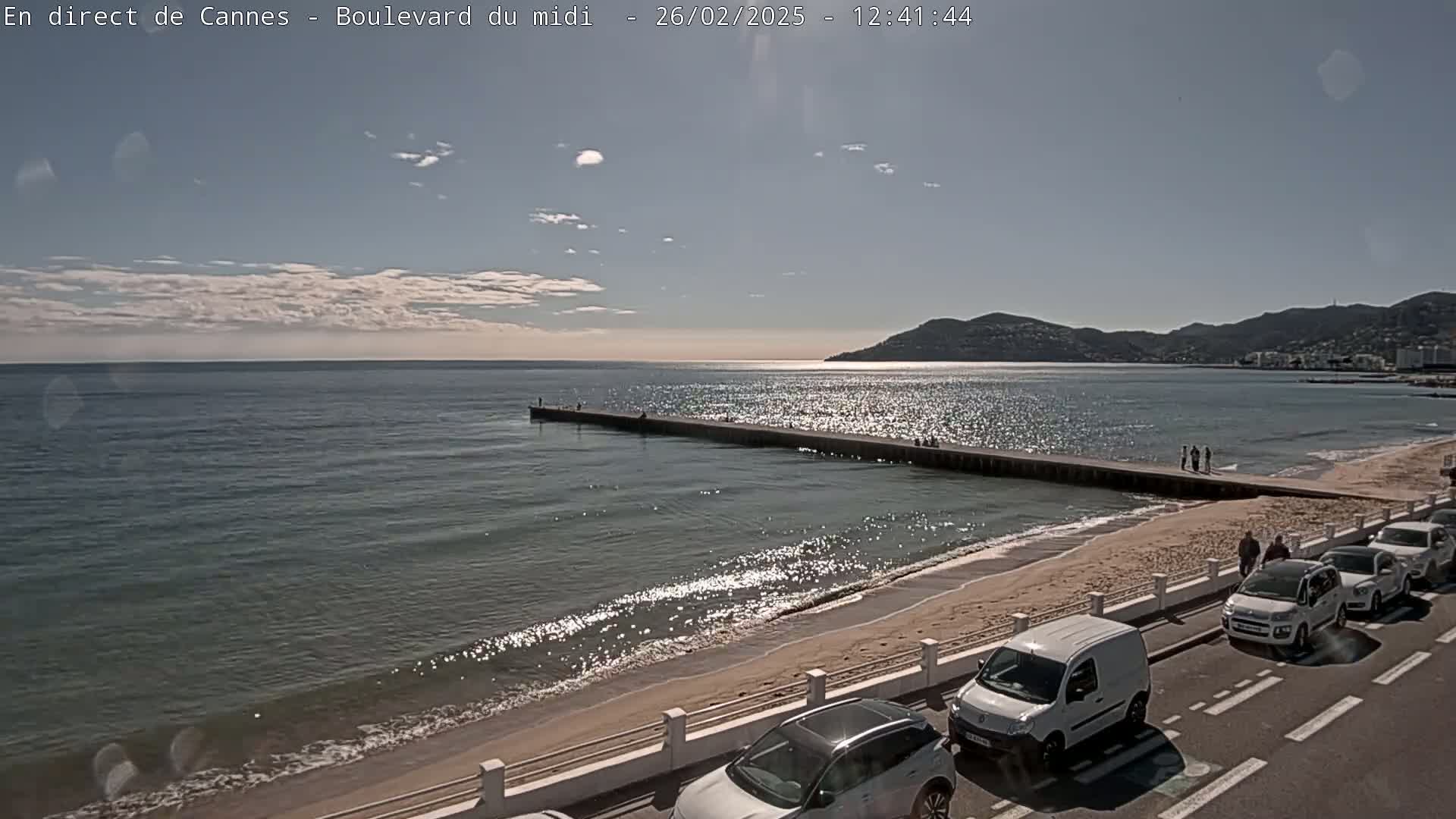 A sandy beach with calm, sparkling water, a long pier, several cars parked along the road, and a partly cloudy sky.