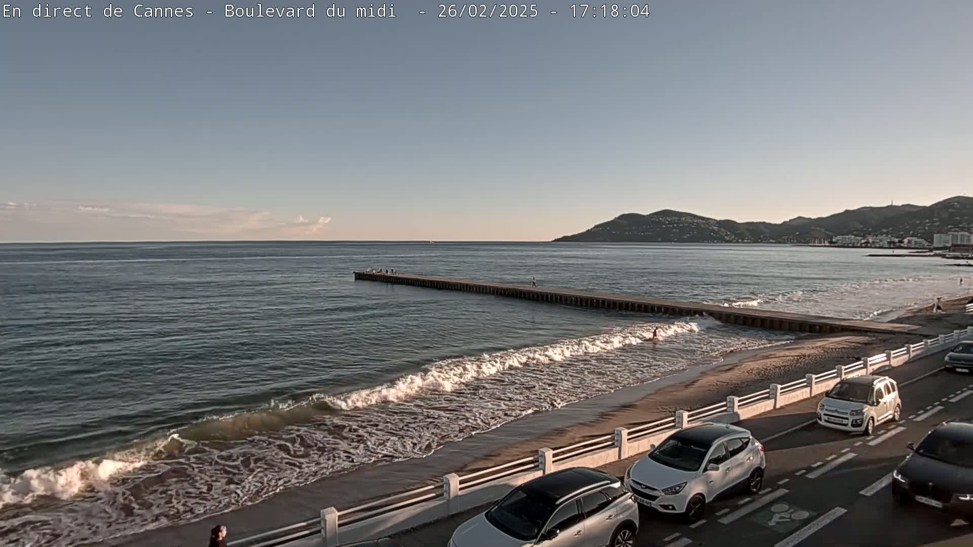 A sandy beach with gentle waves laps against a long pier extending into a calm sea under a mostly clear sky, with cars parked along a road beside it.