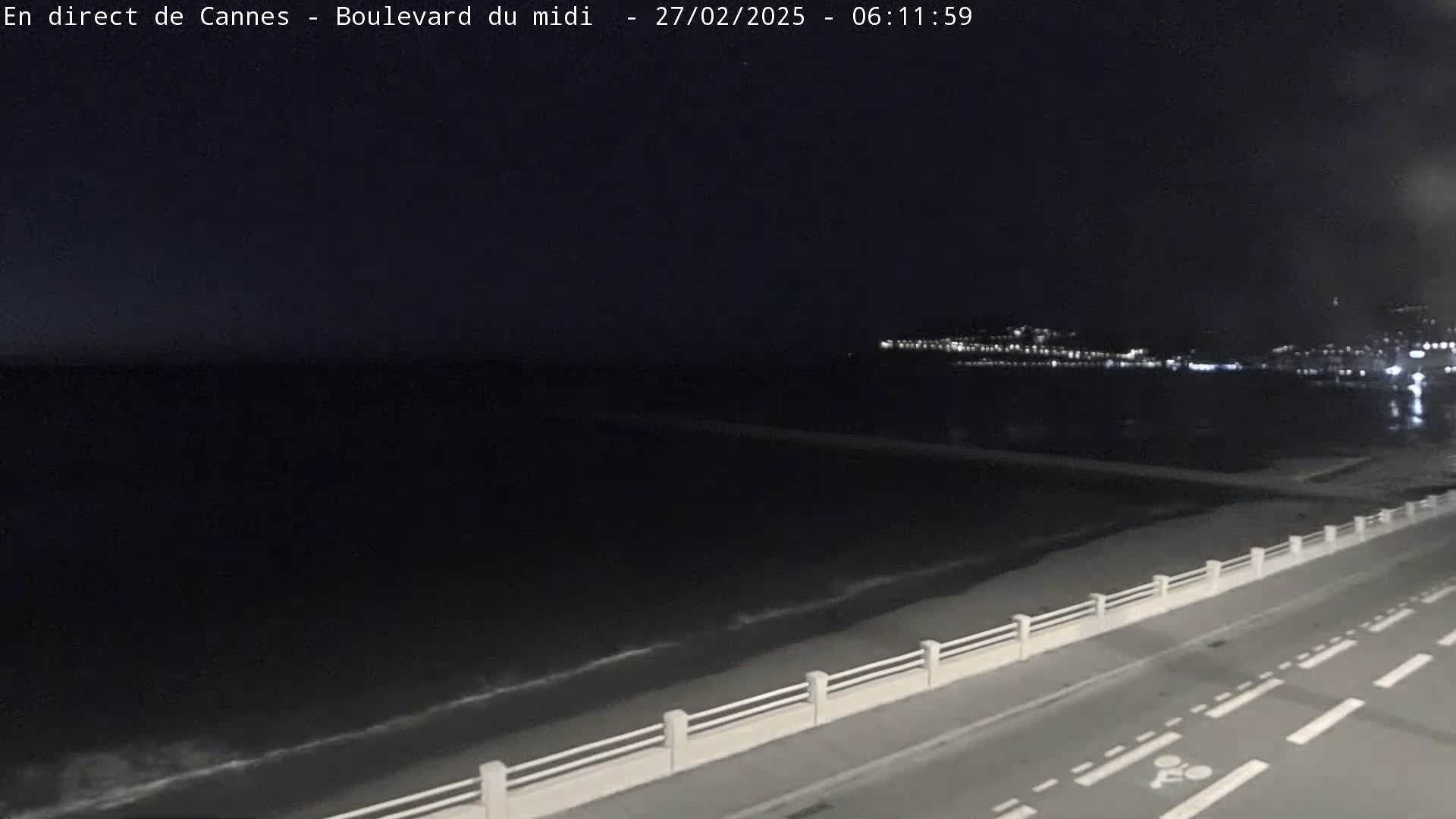 A dark nighttime scene shows a calm sea with small waves lapping a shore with a road and low wall beside it, and distant city lights visible across the water.