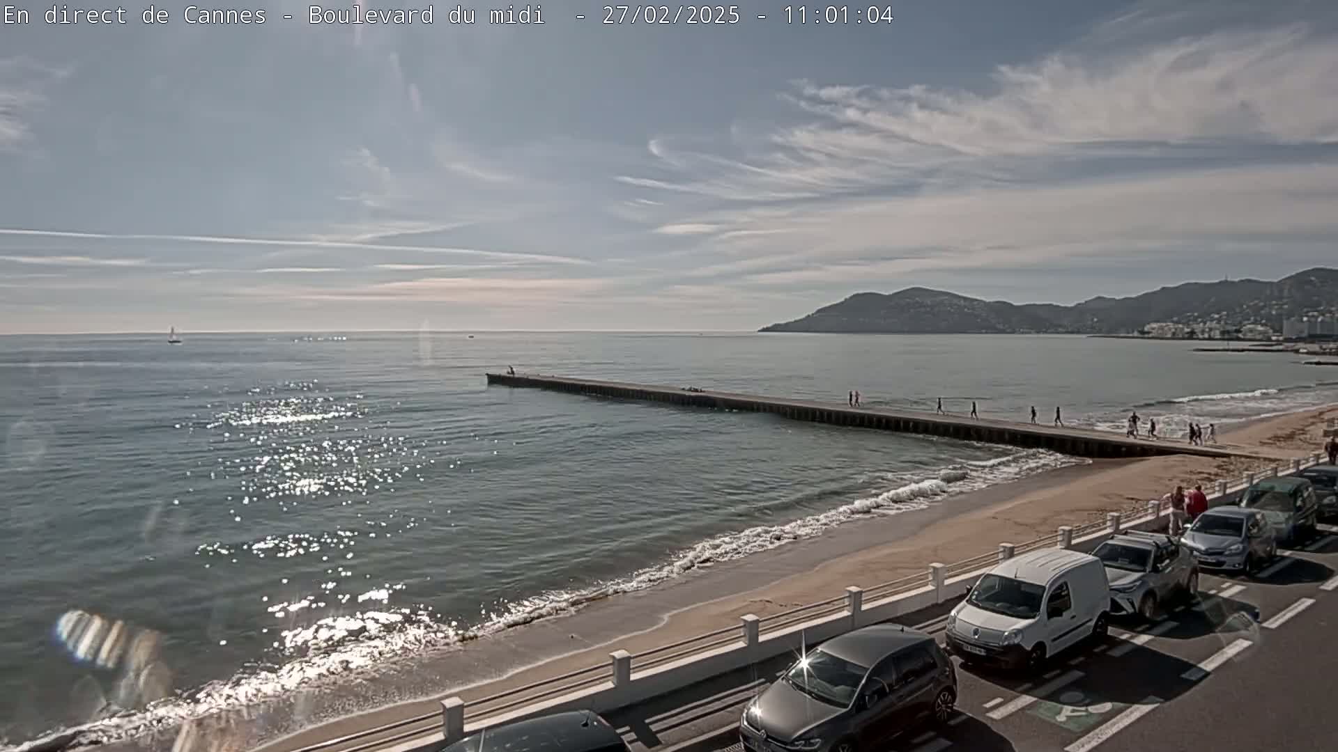 A sandy beach with a long pier extending into a calm, sunlit sea, backed by a line of parked cars and hills in the distance.