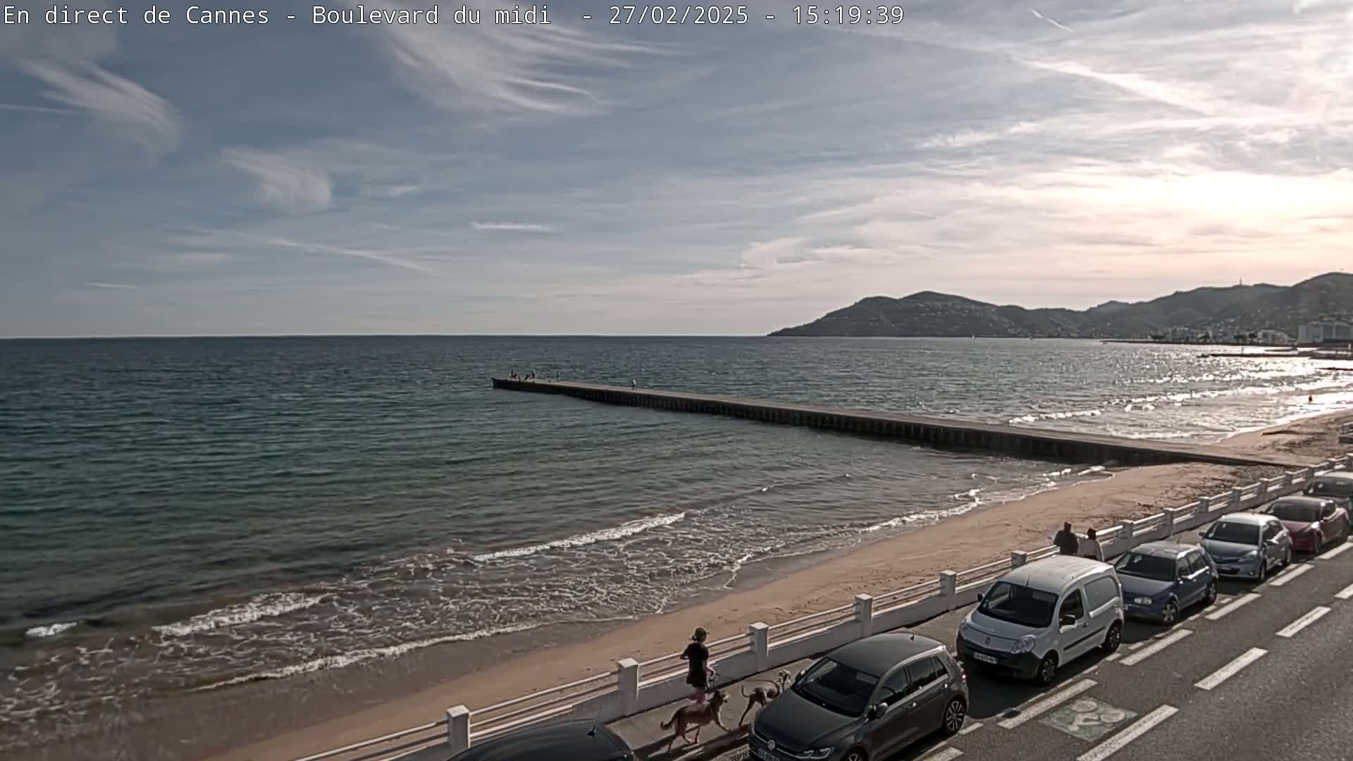 A sandy beach with gentle waves, a long pier extending into a calm sea, cars parked along a road overlooking the water, and a few people walking on the beach under a partly cloudy sky.